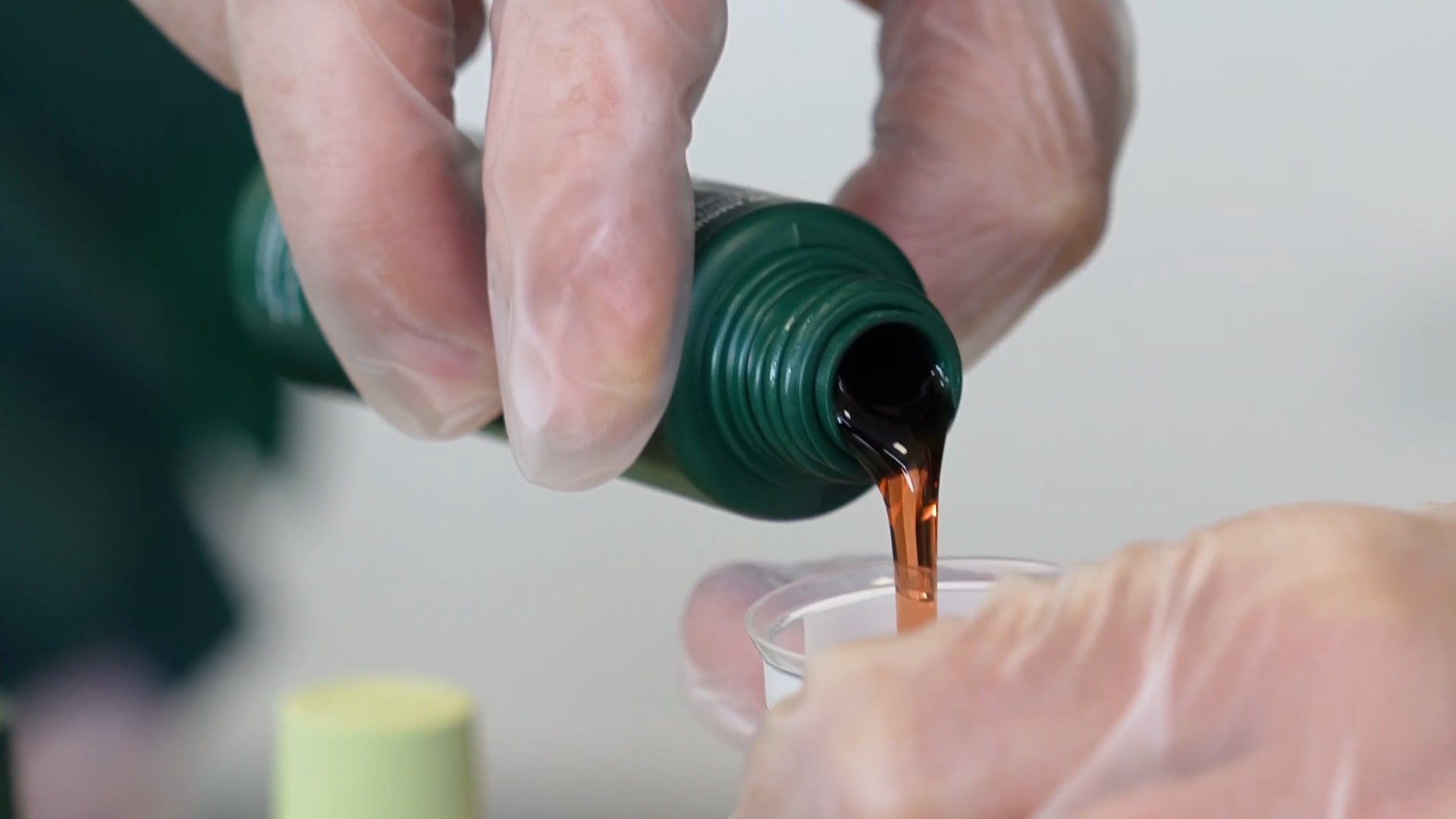 Primo piano di una mano che versa il contenuto di un flacone di colorazione permanente per capelli in un misurino dosatore, su sfondo bianco neutro